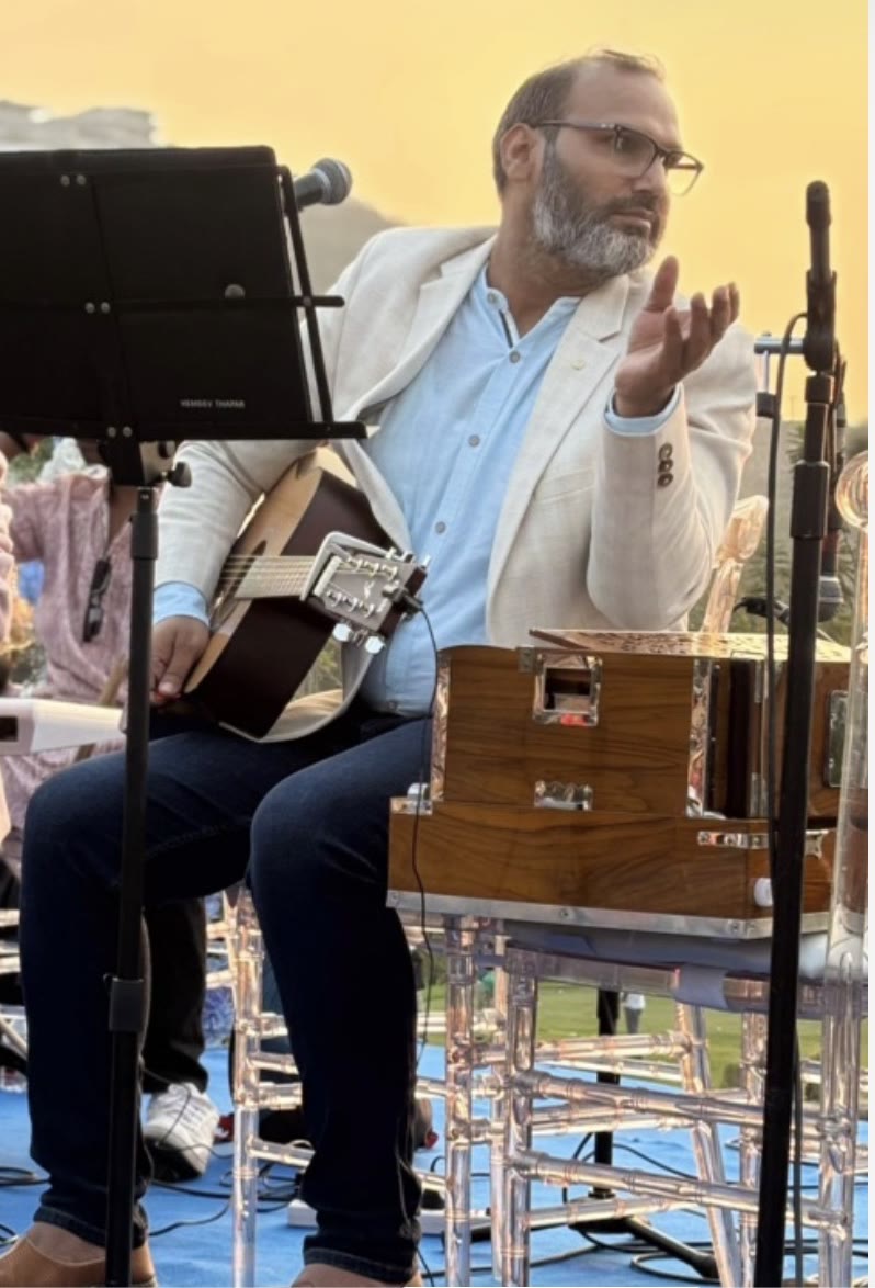 Hemant Acharya at golden hour with a guitar on his lap and a harmonium beside him, one hand raised mid-phrase as he sings